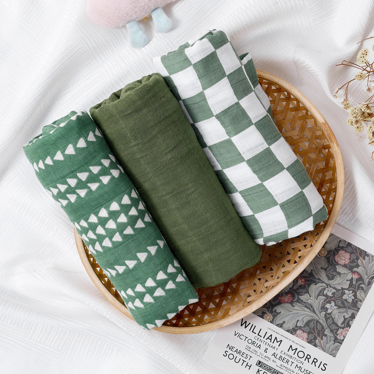 Set of three green muslin baby swaddle blankets with different patterns on a wicker tray
