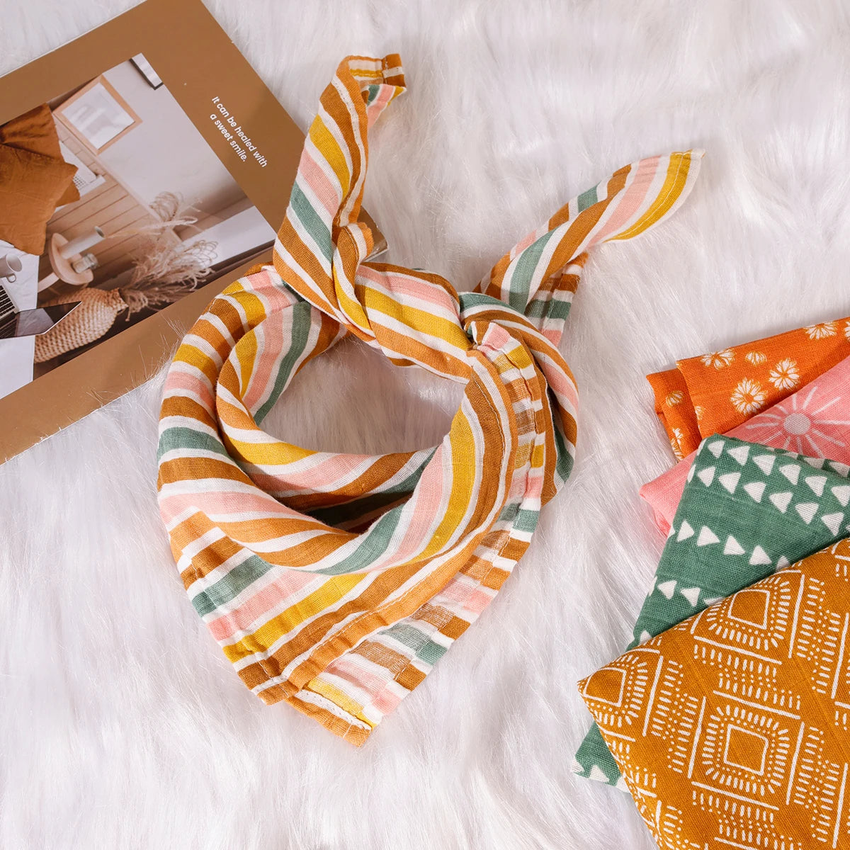 Striped muslin baby bandana bib on soft white fur, with colorful patterned cloths nearby