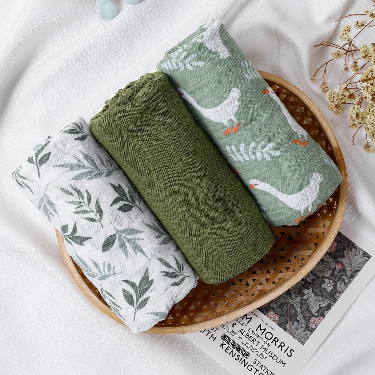 Three rolled muslin baby swaddle blankets with leaf and goose patterns on a bamboo tray