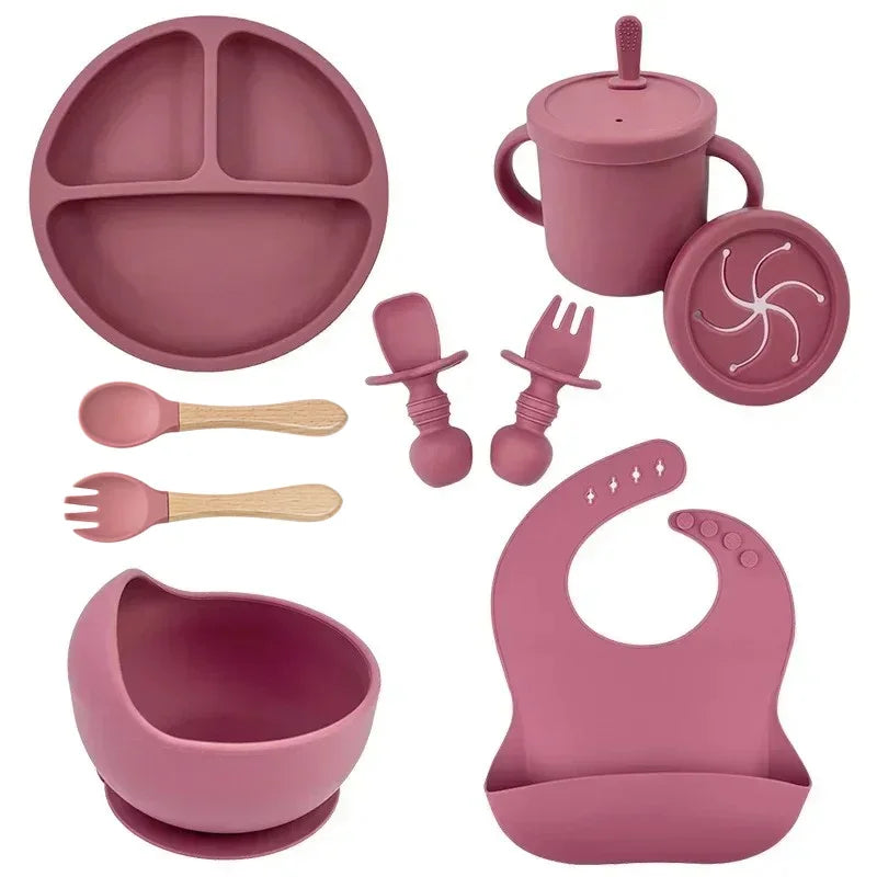 Pink silicone baby feeding set with bib, divided plate, bowl, cup, utensils, and snack lid.