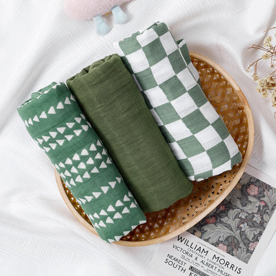 Set of three green muslin baby swaddle blankets with different patterns on a wicker tray