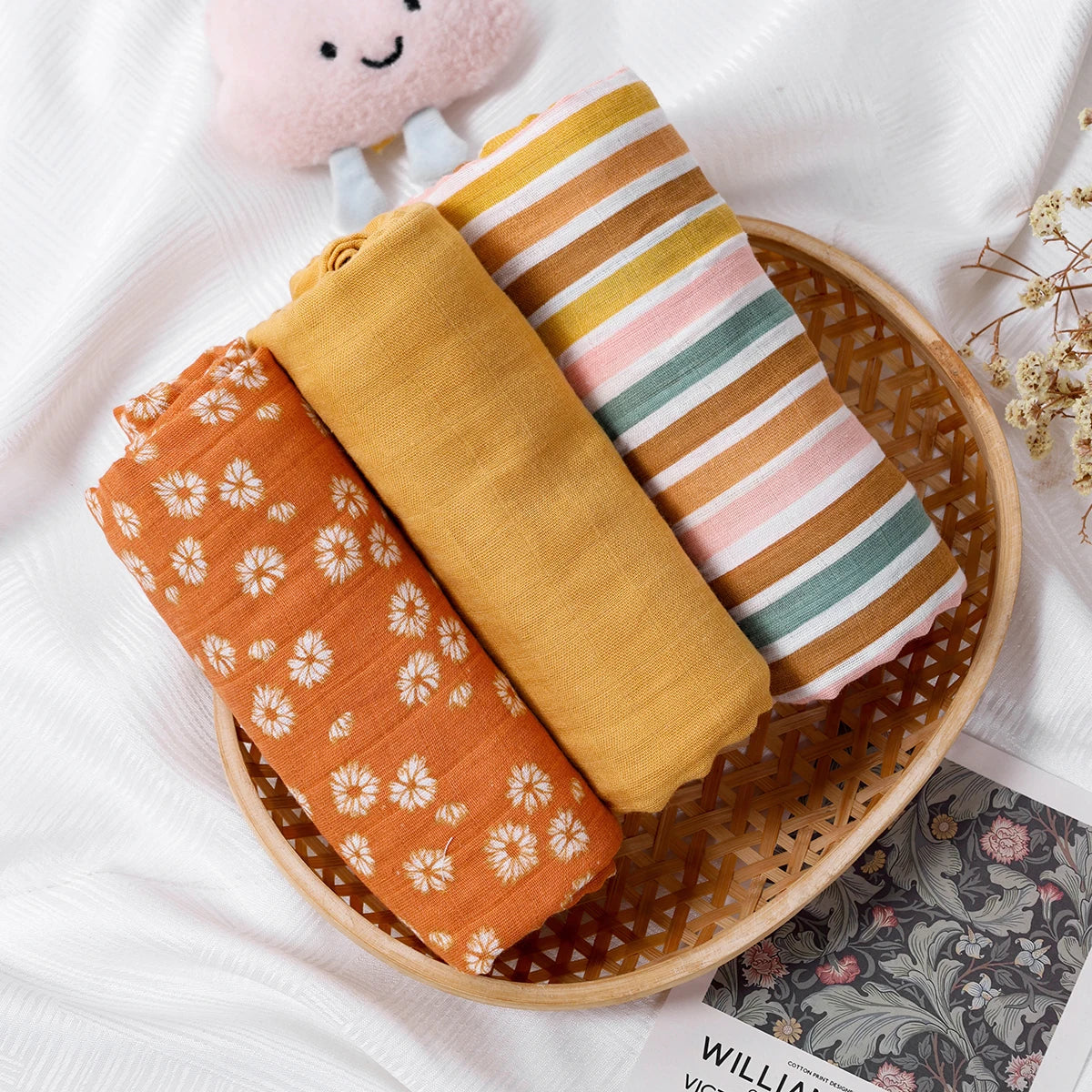 Three rolled baby muslin blankets, floral, solid mustard, and striped, in a basket on white fabric.