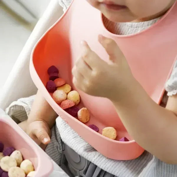 Baby wearing a pink silicone bib, holding colorful snacks during mealtime