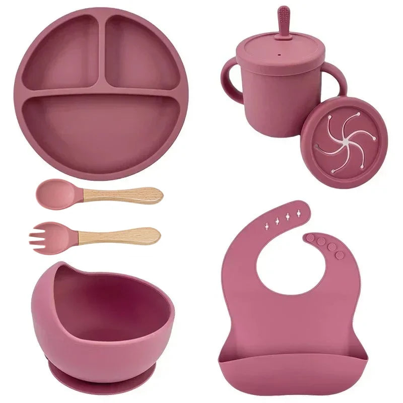 Mauve silicone baby feeding set with divided plate, sippy cup, bowl, spoon, fork, and bib.